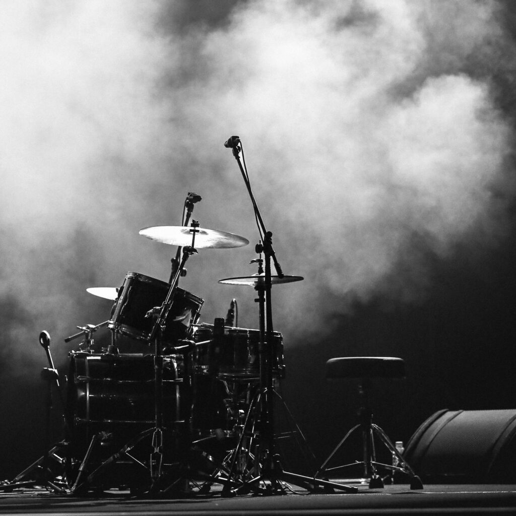 A dramatic black and white image of a drum set on stage with spotlights and smoke